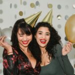 Young cheerful friends in shiny birthday caps with silver and golden balloons among shimmering confetti