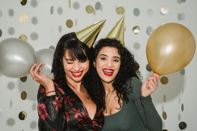 Young cheerful friends in shiny birthday caps with silver and golden balloons among shimmering confetti